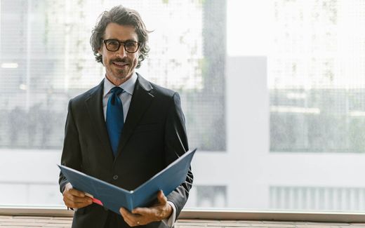 A smiling executive in a suit stands holding an open folder in a bright, window-lit space