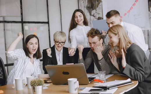 Six colleagues gather around a laptop in a bright office, pumping fists and cheering together