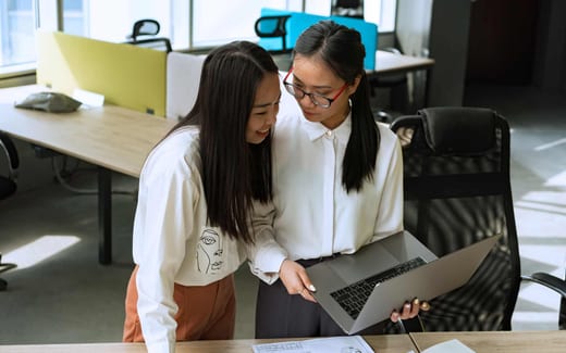 Two colleagues in a bright open office review a floor-plan drawing on a laptop and printed sheets