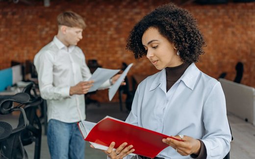 A woman holds an open red folder and reviews its contents while a colleague looks over papers behind her in a brick-walled office
