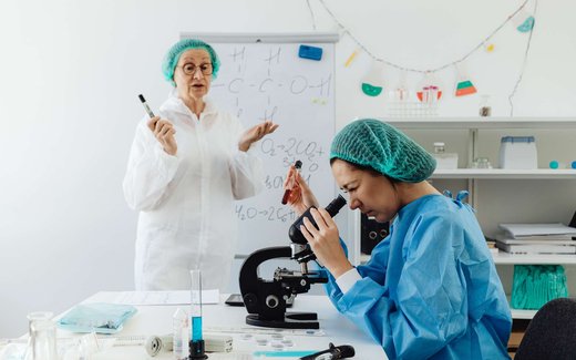 A researcher in a lab coat gestures toward a whiteboard of chemical formulas while a colleague examines a sample under a microscope