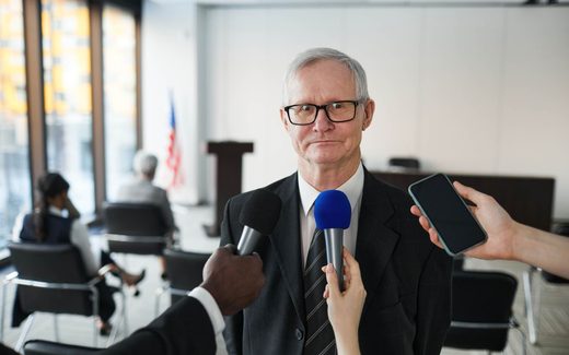 An older man in glasses answers questions as microphones and a phone are held toward him