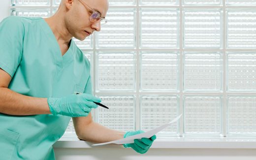 A gloved clinician in scrubs reviews a printed document by a bright glass-block window