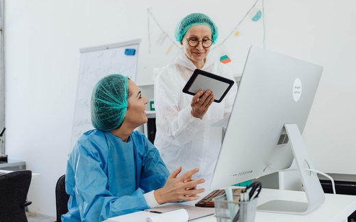 Two colleagues in surgical caps and protective gowns review notes on a tablet in a bright laboratory office