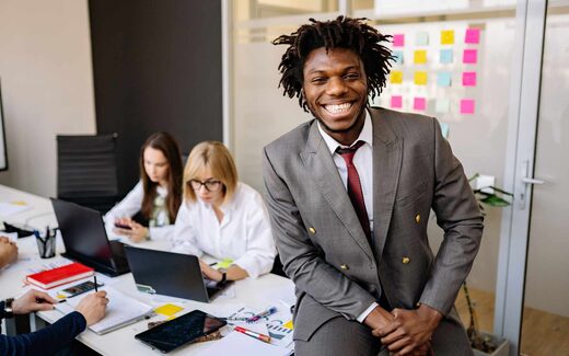 A smiling executive in a grey suit sits on the edge of a desk while two colleagues work at laptops behind him in a bright office