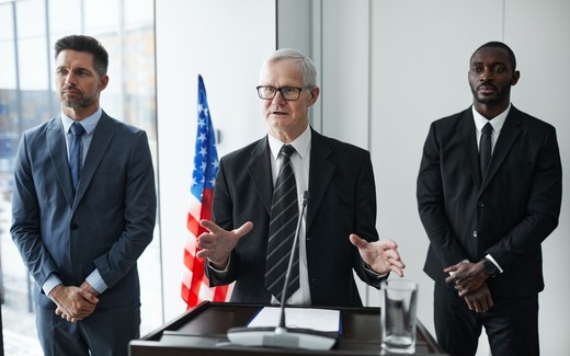 Three men in business suits stand at a lectern with a U.S. flag behind them, one gesturing as he speaks