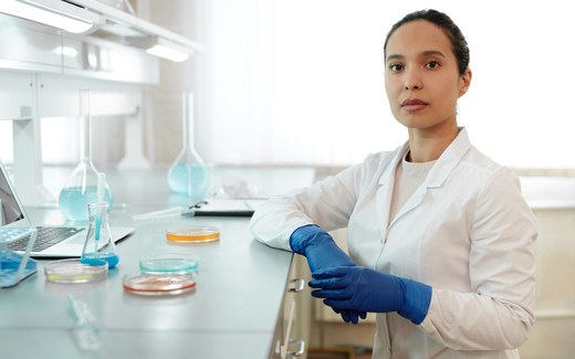 A scientist in a white lab coat and blue nitrile gloves stands at a laboratory bench beside petri dishes and glass flasks