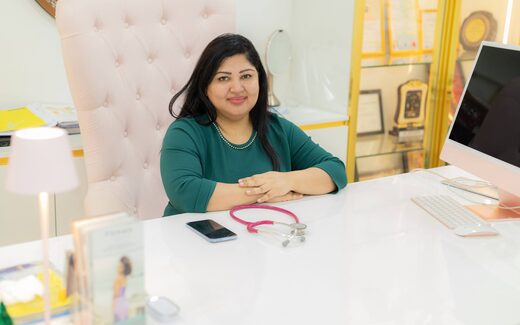A physician sits at a bright, tidy desk with a stethoscope, a phone and a desktop computer
