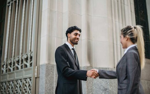 Two professionals in suits shake hands in front of a stone-columned government building facade