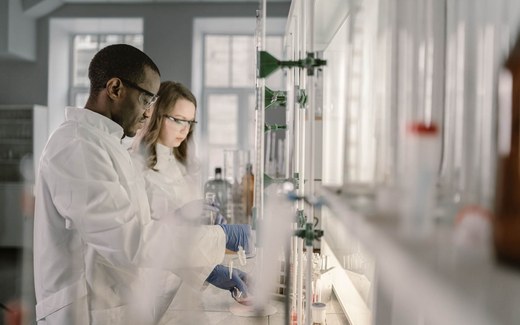 Two lab scientists in white coats test glassware samples on a bright laboratory bench