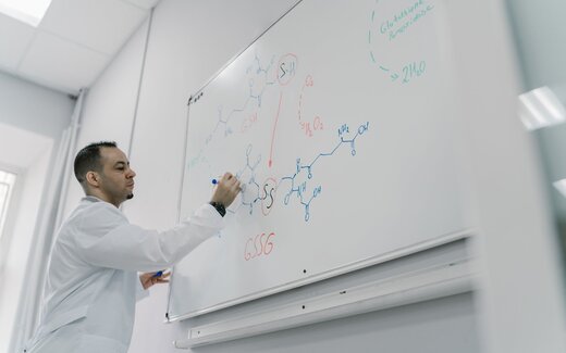 A scientist in a lab coat diagrams a molecular structure on a whiteboard
