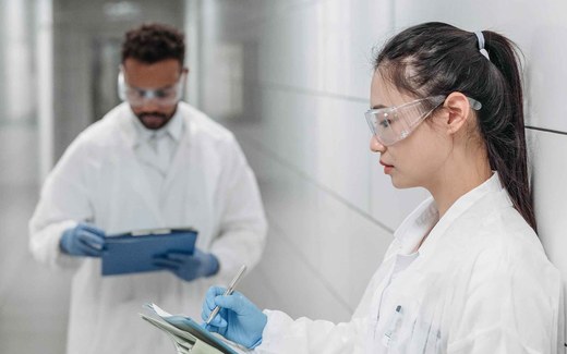 A scientist in a white lab coat and safety glasses reviews handwritten notes on a clipboard in a laboratory corridor, with a colleague behind her