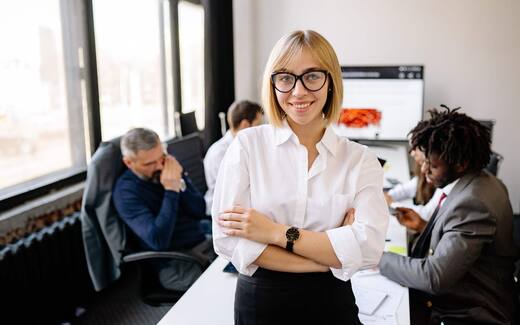 A business leader stands in an office with colleagues working in the background