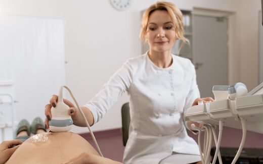 A clinician in a white coat holds an ultrasound transducer against a patient while reading a nearby ultrasound machine display