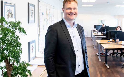 A man in a blazer smiles in a bright office hallway, with colleagues working at standing desks behind him