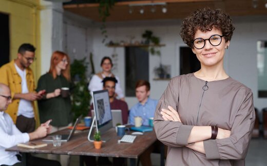 A confident executive stands with arms crossed in a loft office while colleagues confer behind her, standing in for Verastem Oncology's financing announcement