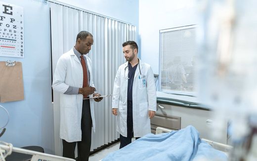 Two physicians in white coats review a chart on a clipboard together in a hospital exam room