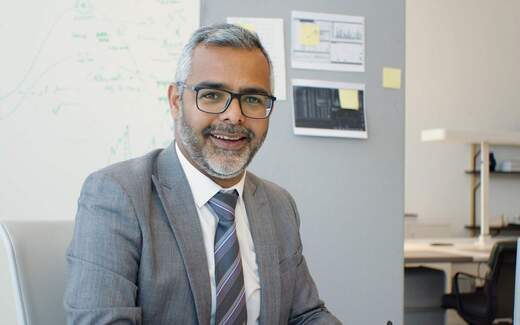A businessman in glasses and a grey suit smiles in an office, with data charts pinned to the wall behind him
