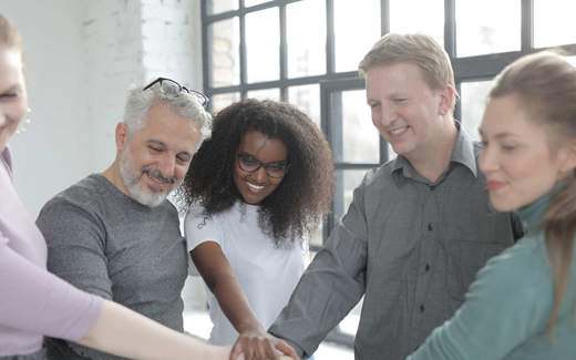 A team of five colleagues put their hands together in the middle of a bright, window-lit room, celebrating together
