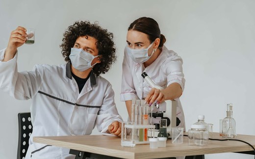 Two masked researchers in a lab examine a beaker of green solution and adjust a microscope and test tubes on a workbench