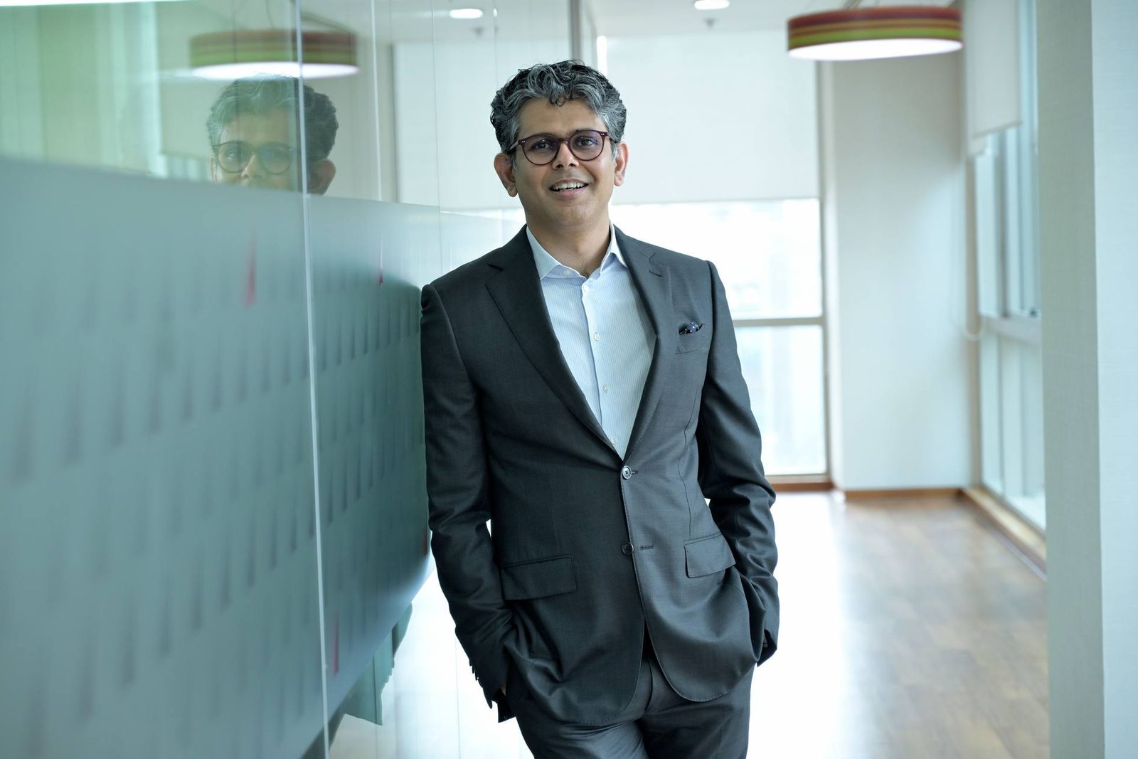 A confident executive in a gray suit stands in a bright glass-walled office corridor