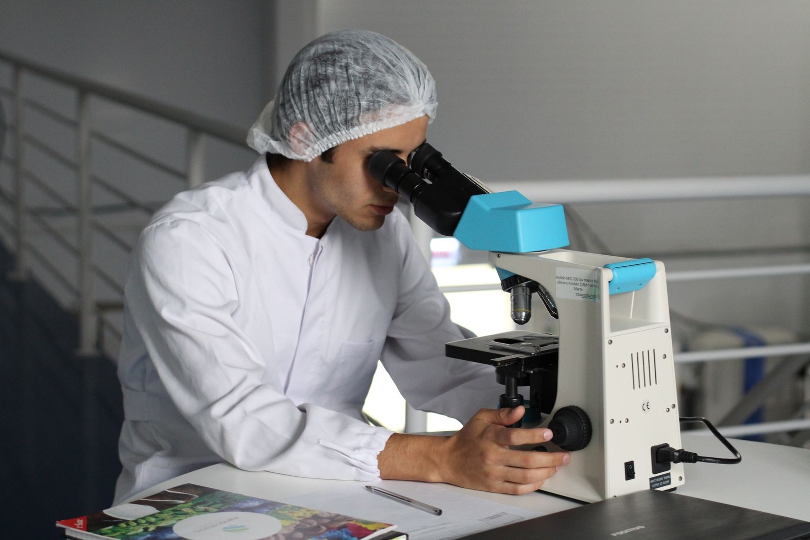 A researcher in a hairnet and lab coat looks through a benchtop microscope while adjusting its focus knob