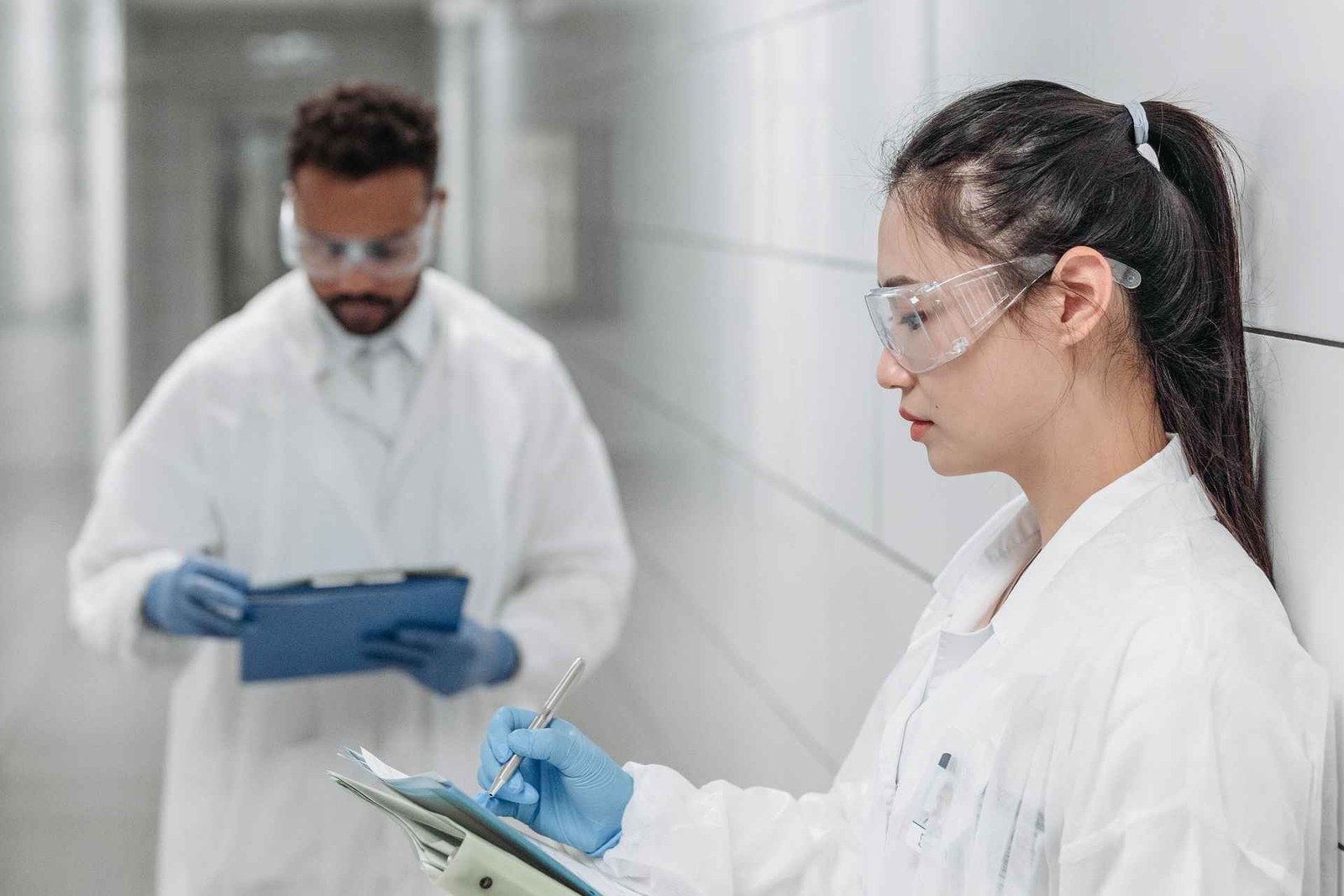 A scientist in a white lab coat and safety glasses reviews handwritten notes on a clipboard in a laboratory corridor, with a colleague behind her