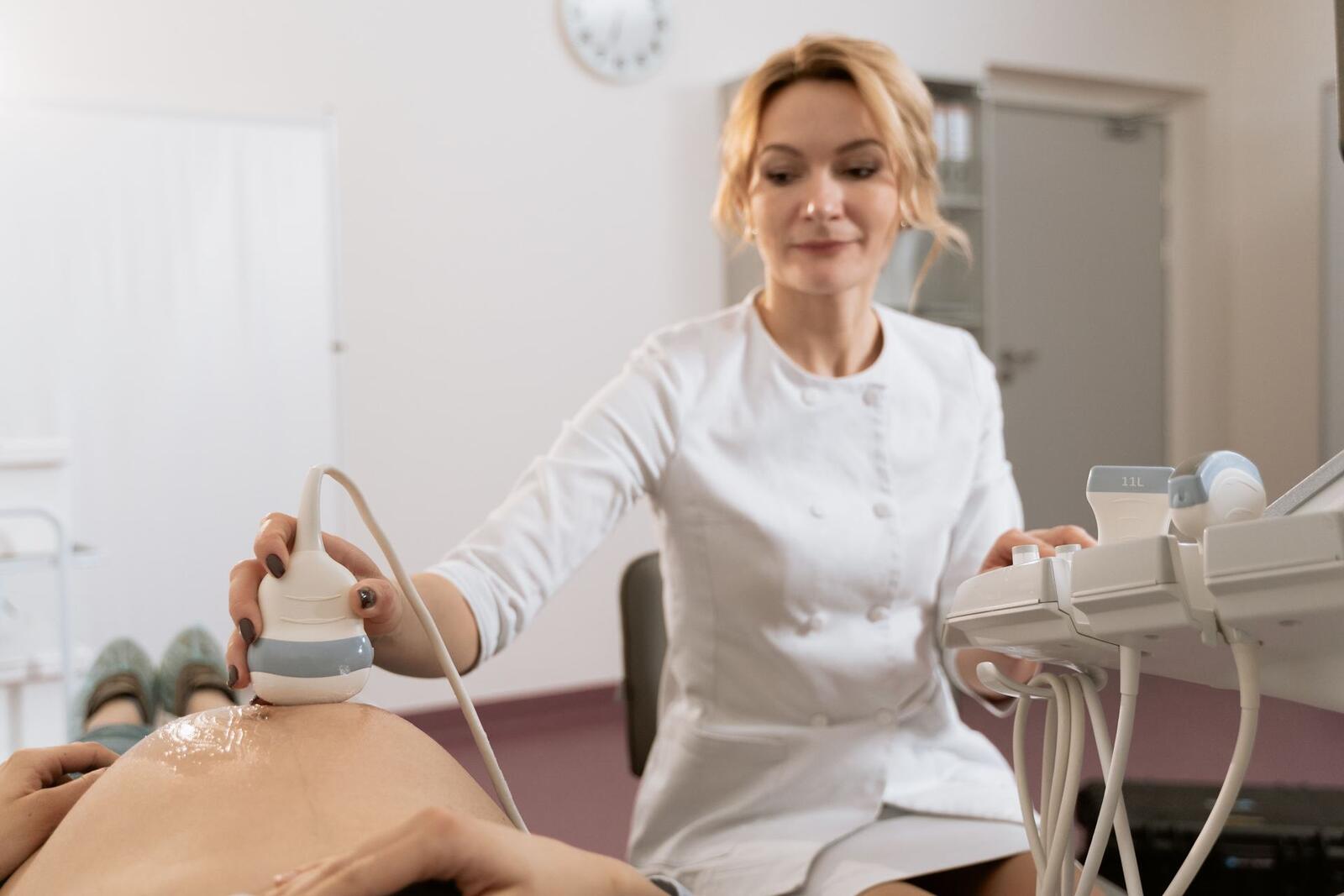 A clinician in a white coat holds an ultrasound transducer against a patient while reading a nearby ultrasound machine display