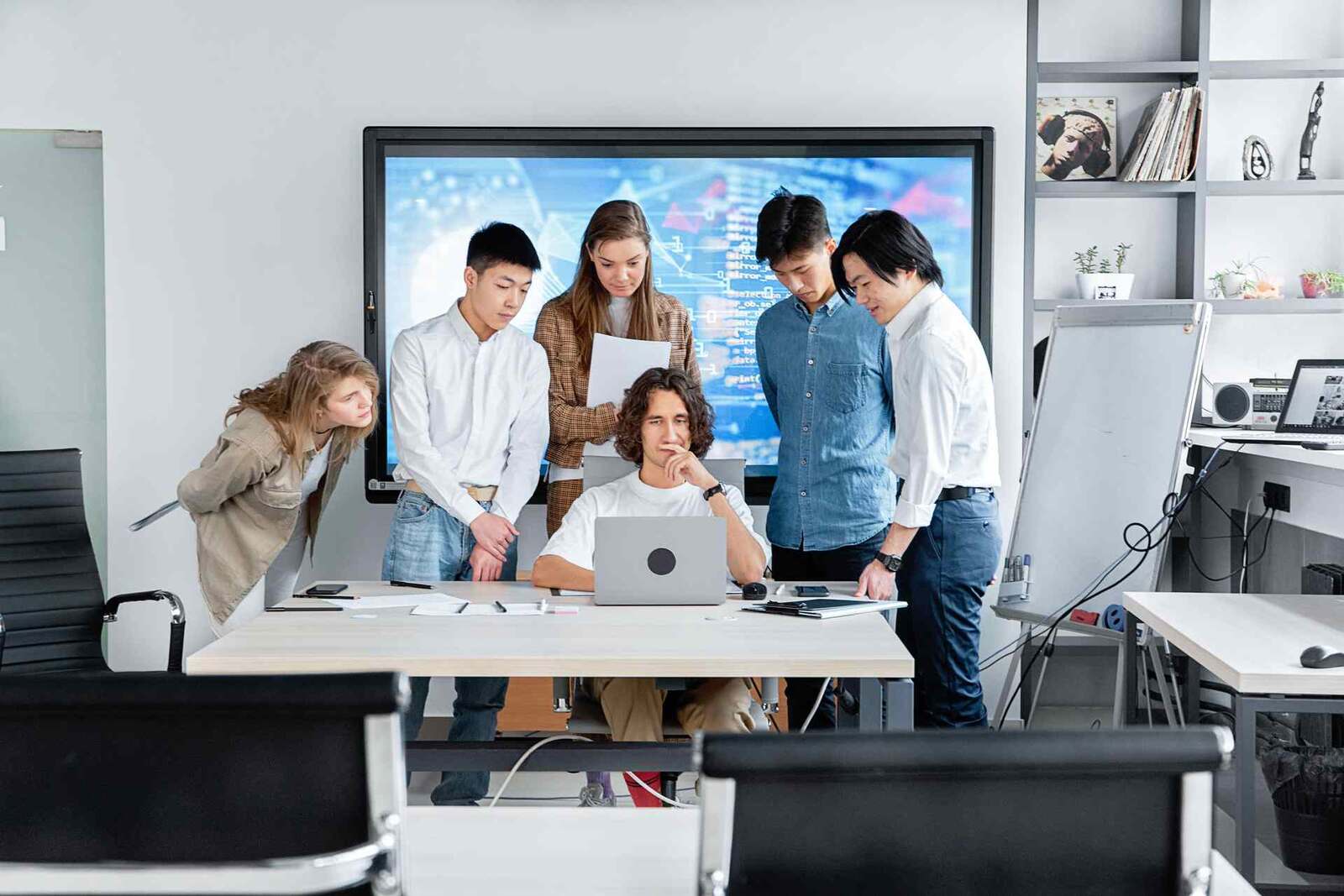 A product team gathers around a laptop and a wall display showing data during a design review