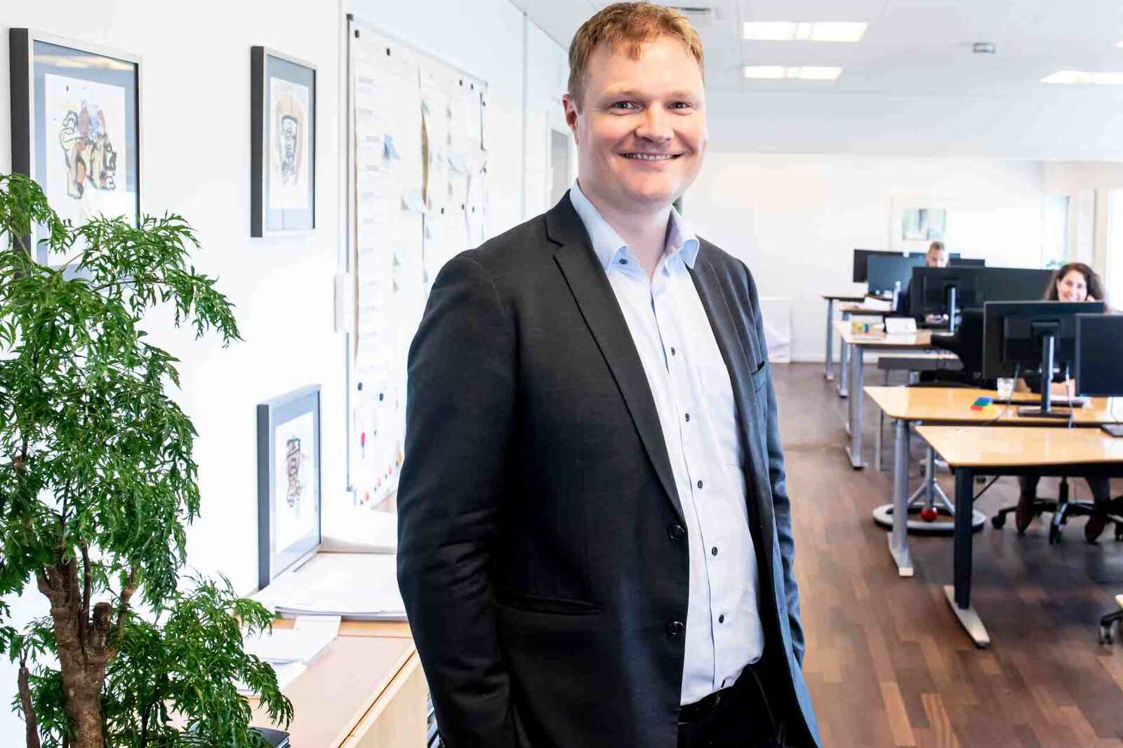 A man in a blazer smiles in a bright office hallway, with colleagues working at standing desks behind him