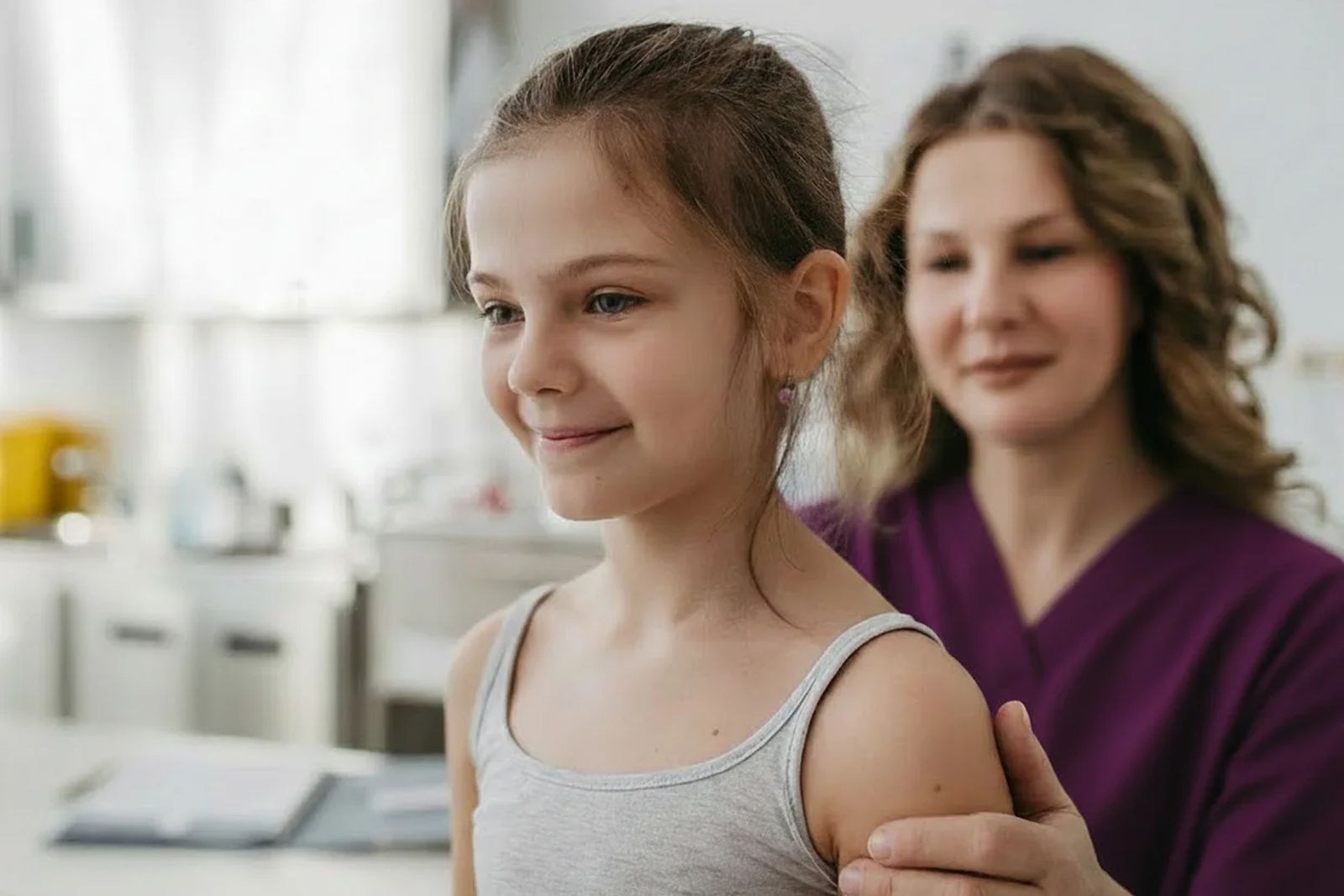 A smiling girl in a grey tank top stands in an exam room while a clinician in purple scrubs rests a hand on her shoulder, from VB Spine's website