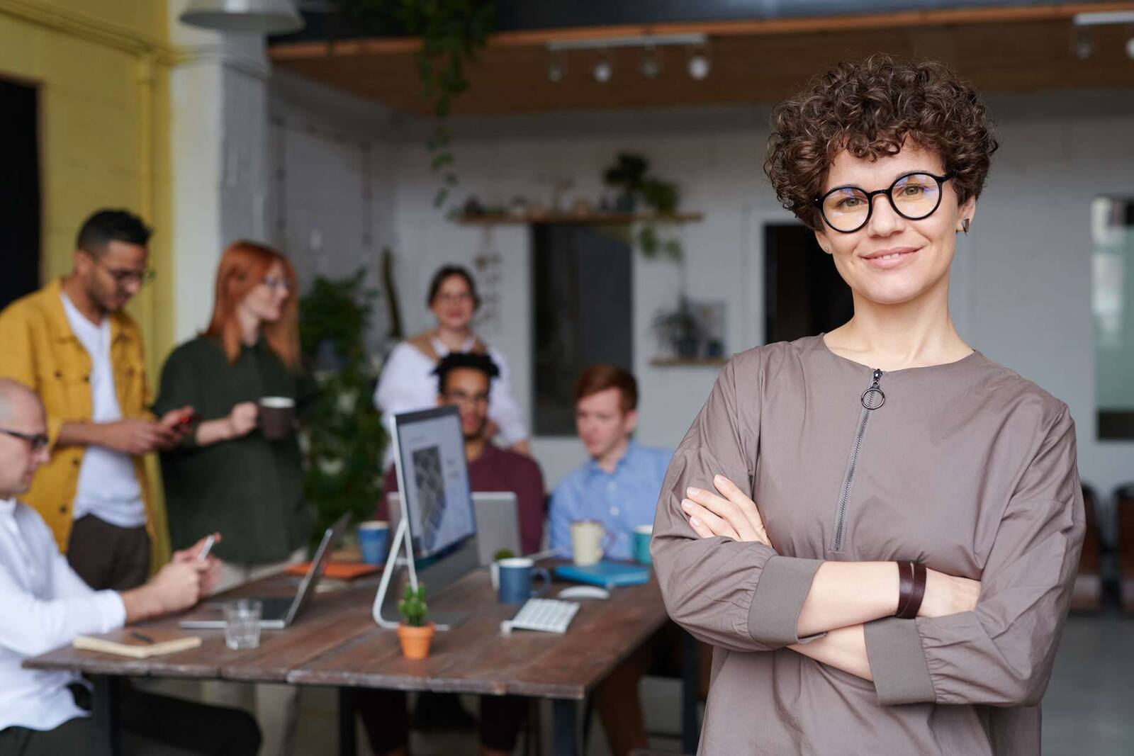 A confident executive stands with arms crossed in a loft office while colleagues confer at a table behind her