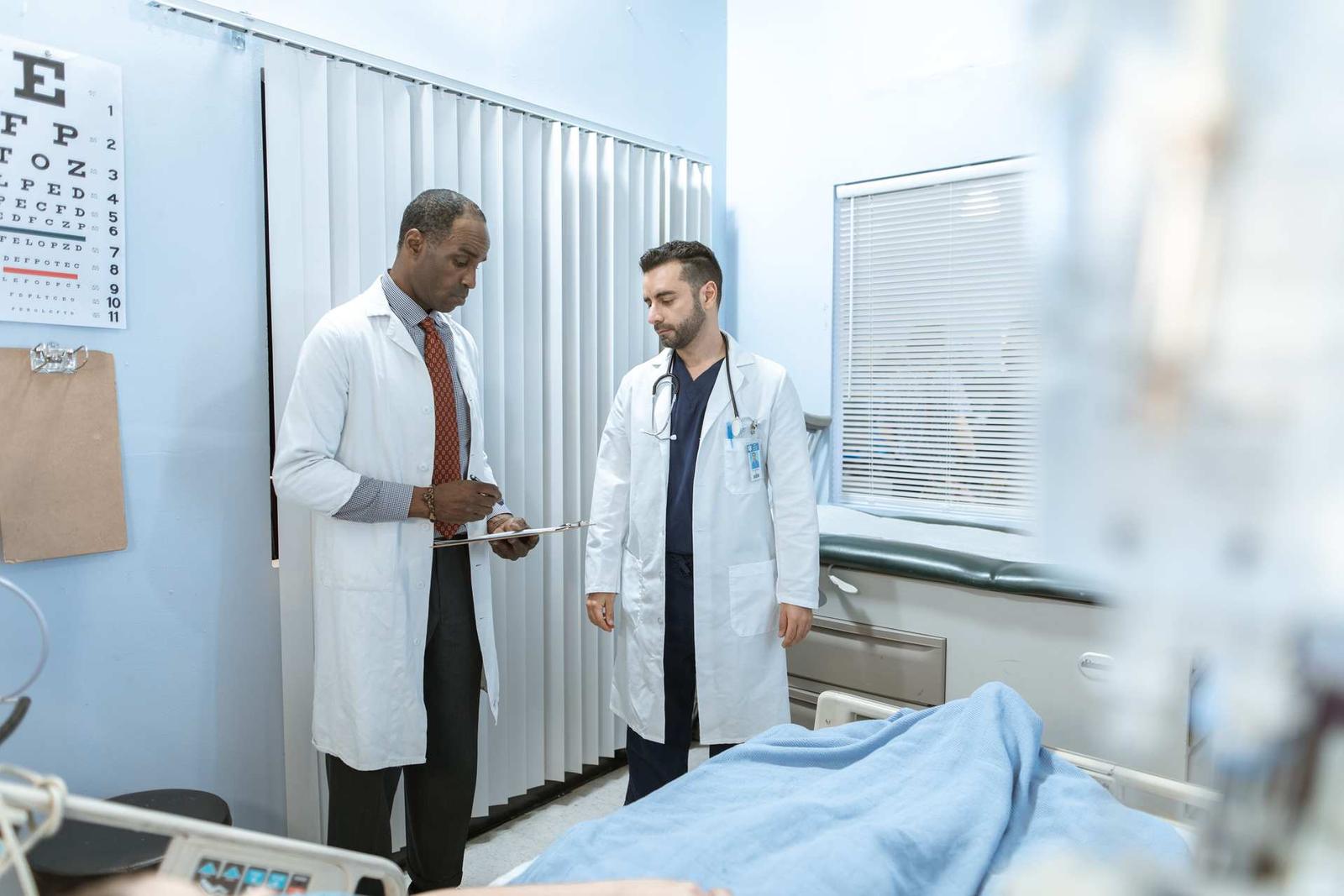Two physicians in white coats review a chart on a clipboard together in a hospital exam room