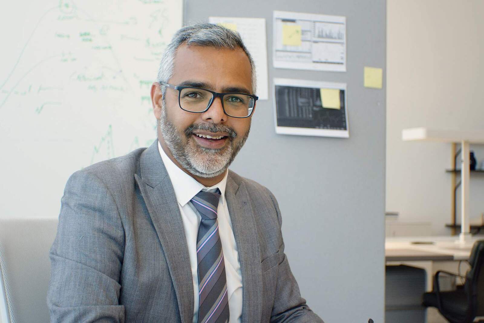 A businessman in glasses and a grey suit smiles in an office, with data charts pinned to the wall behind him