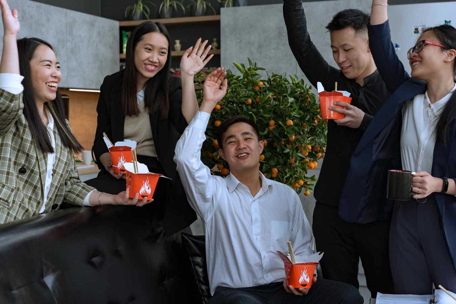 A small office team raising their hands and smiling around takeout containers, a generic stand-in for an approval-day reaction — not Xbrane's own staff or offices