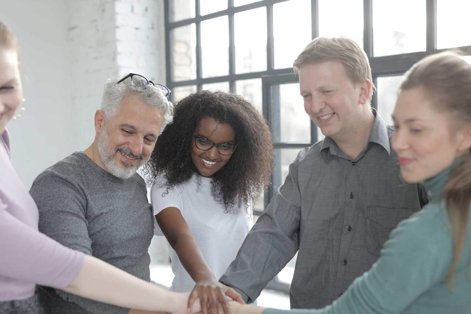 A team of five colleagues put their hands together in the middle of a bright, window-lit room, celebrating together