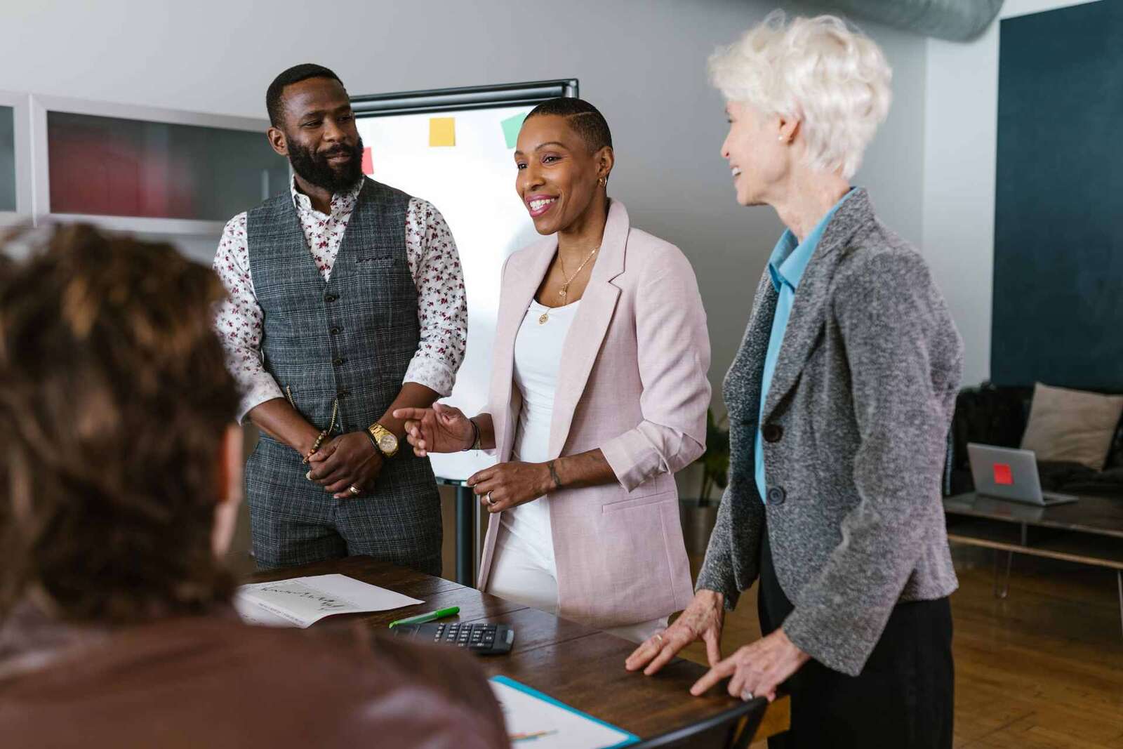 Four colleagues in business attire talking around a conference table in front of a flipchart in a bright office