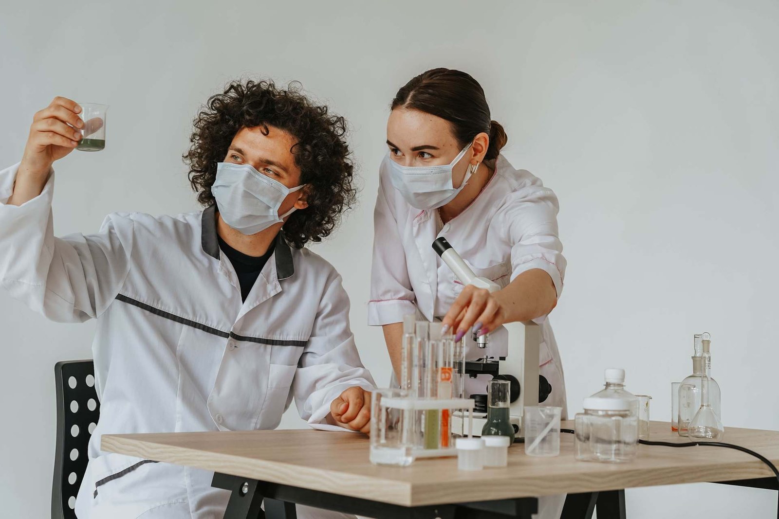 Two masked researchers in a lab examine a beaker of green solution and adjust a microscope and test tubes on a workbench