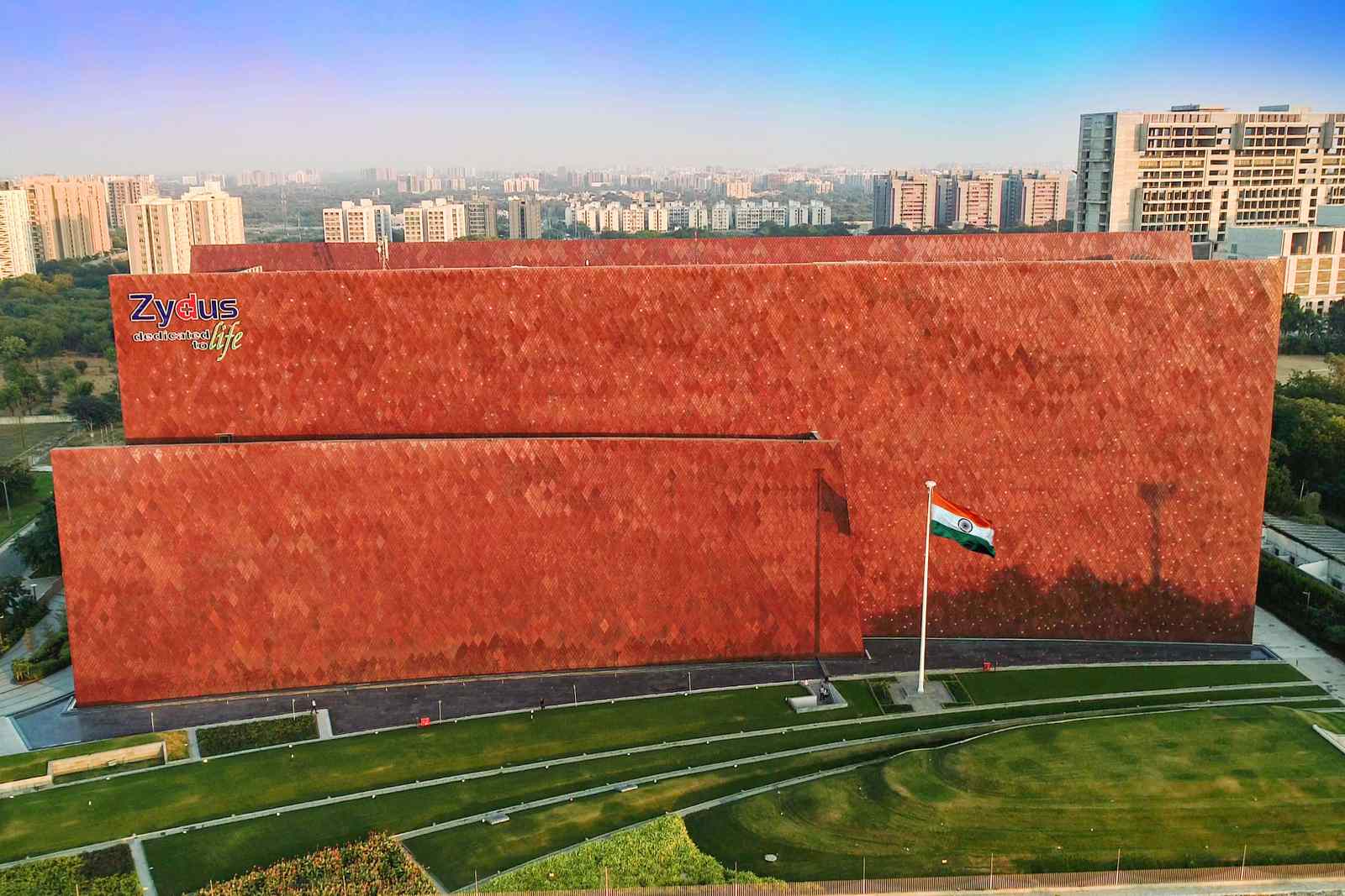 Aerial view of Zydus Corporate Park in Ahmedabad: a long building clad in terracotta diamond-pattern tiles with the Zydus sign at its upper left, an Indian flag on a pole before it and landscaped lawns in front, apartment towers behind
