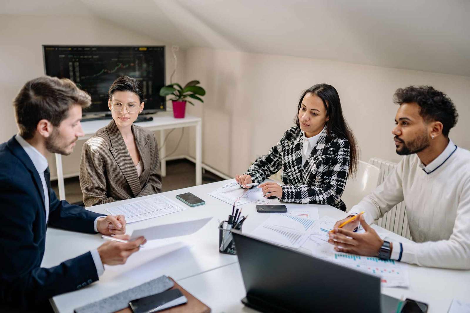Four colleagues in business attire sit and stand around a white conference table covered in printed charts, in a bright meeting room with a wall screen showing a faint data display behind them