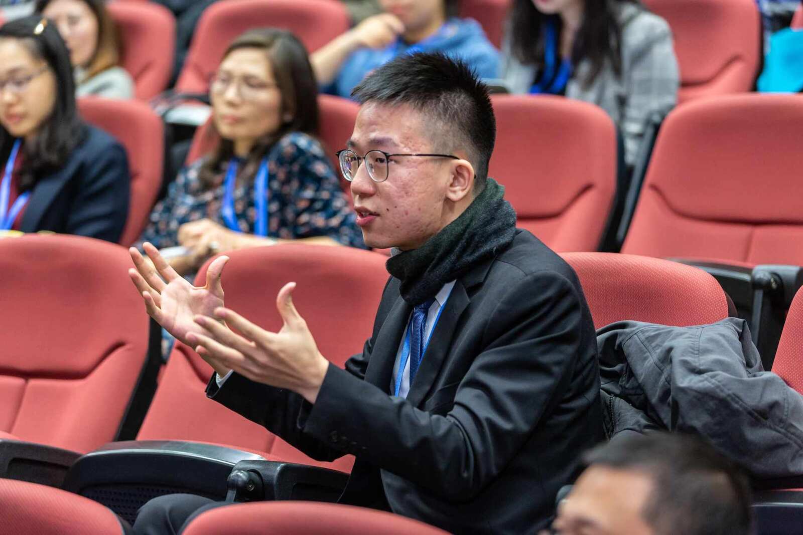 A delegate in a dark suit and lanyard asks a question from the red auditorium seats, hands open mid-sentence, other delegates with lanyards seated behind