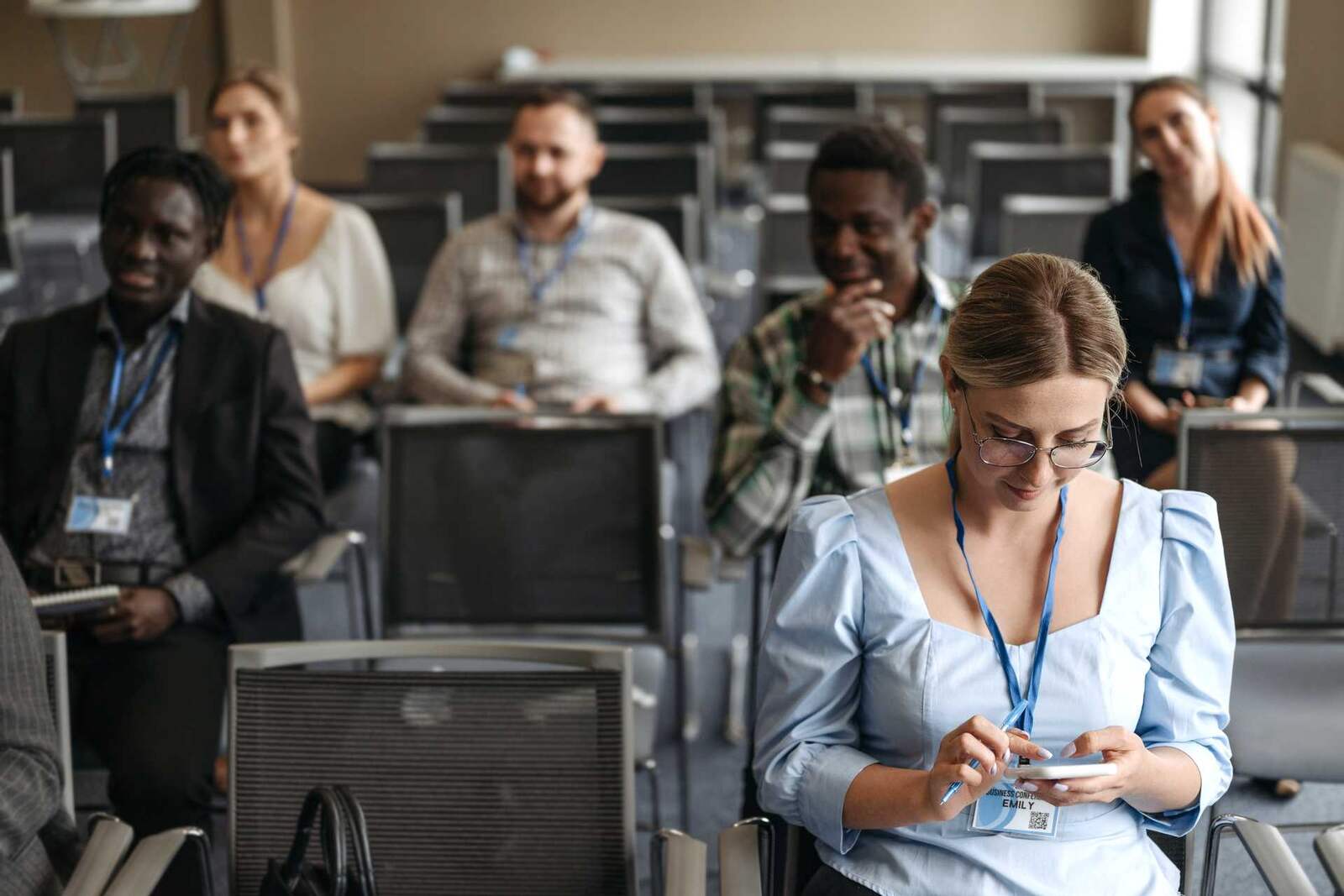 A conference attendee seated in the audience looks down at a phone, attentive to a session in progress