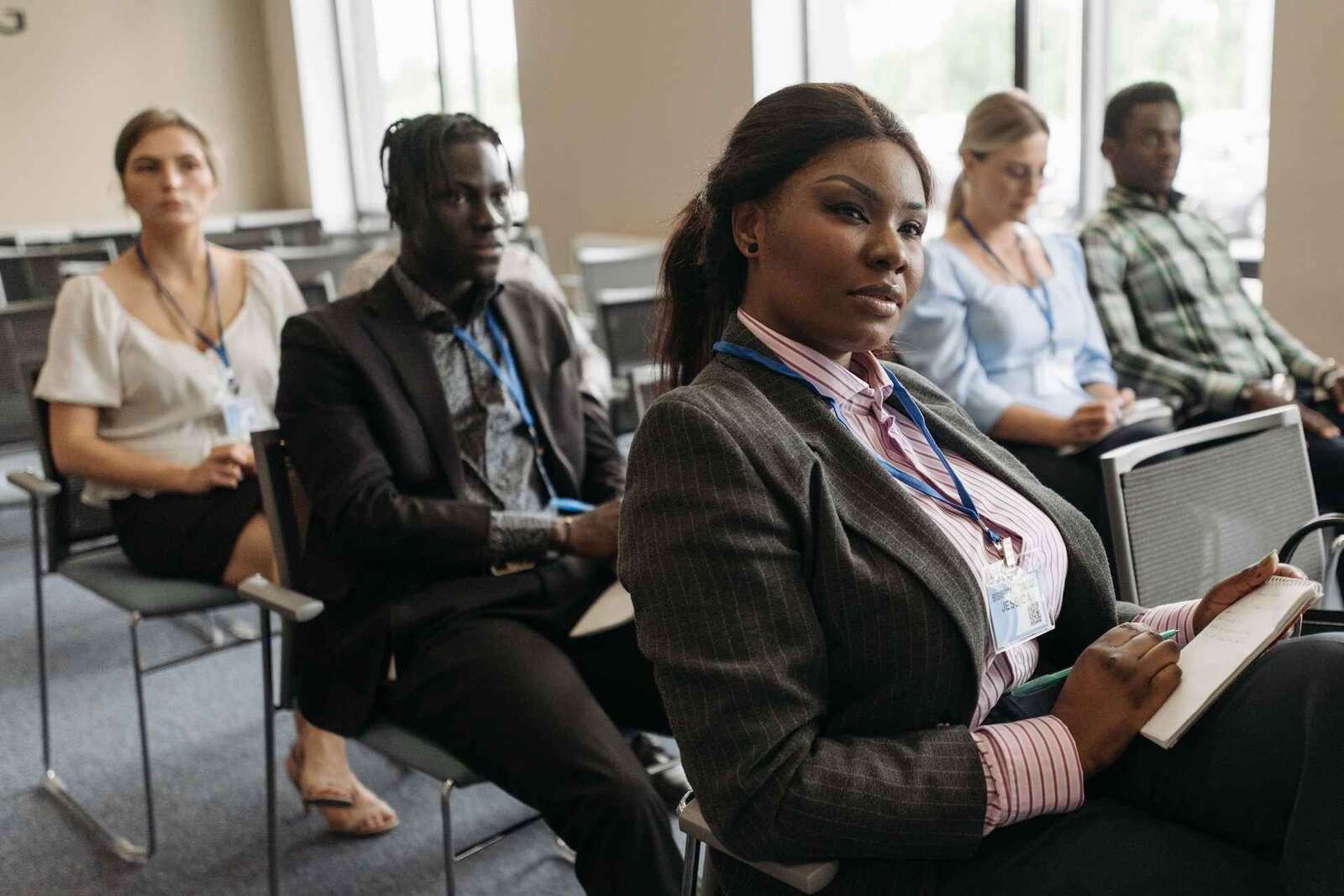 A delegate in a pinstripe blazer with a conference badge sits attentively in the front row, notebook in hand, while colleagues sit around her