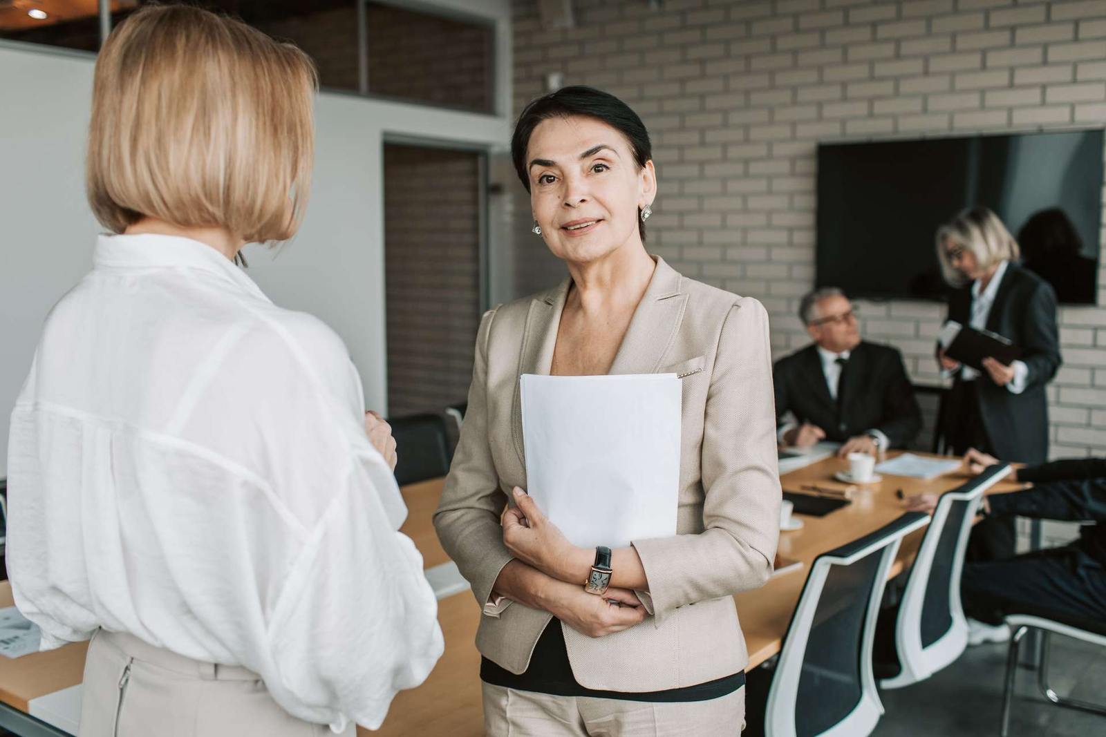 A senior woman in a beige blazer holds papers while talking with a colleague in a boardroom, more attendees seated around a wooden conference table in the background