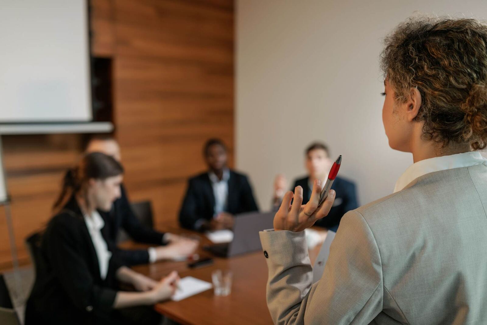 Seen from behind, a presenter in a grey blazer raises a pen while addressing colleagues seated around a boardroom table