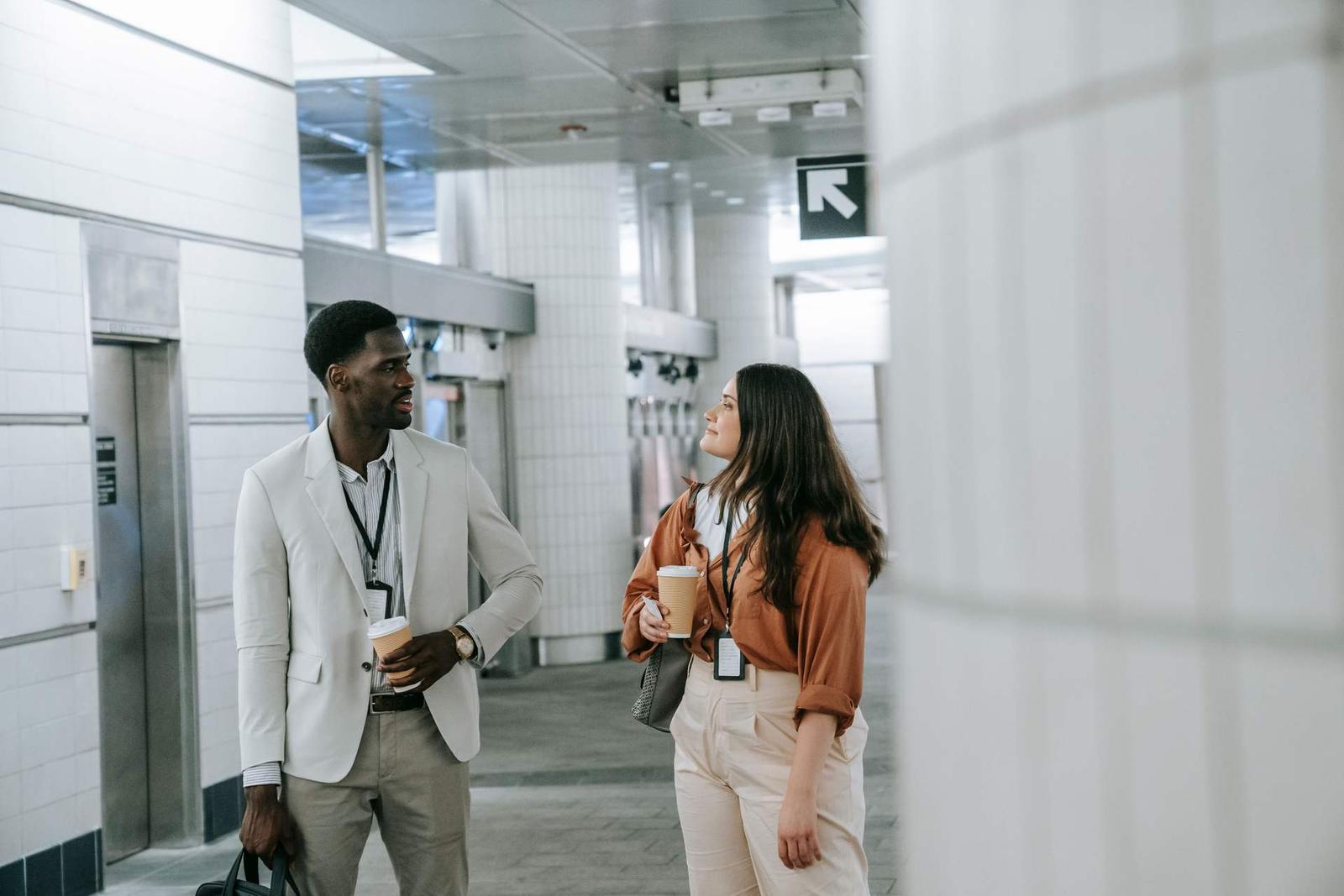 Two colleagues in business attire pause in a bright transit concourse, coffee cups and lanyards in hand, mid-conversation beneath a directional sign