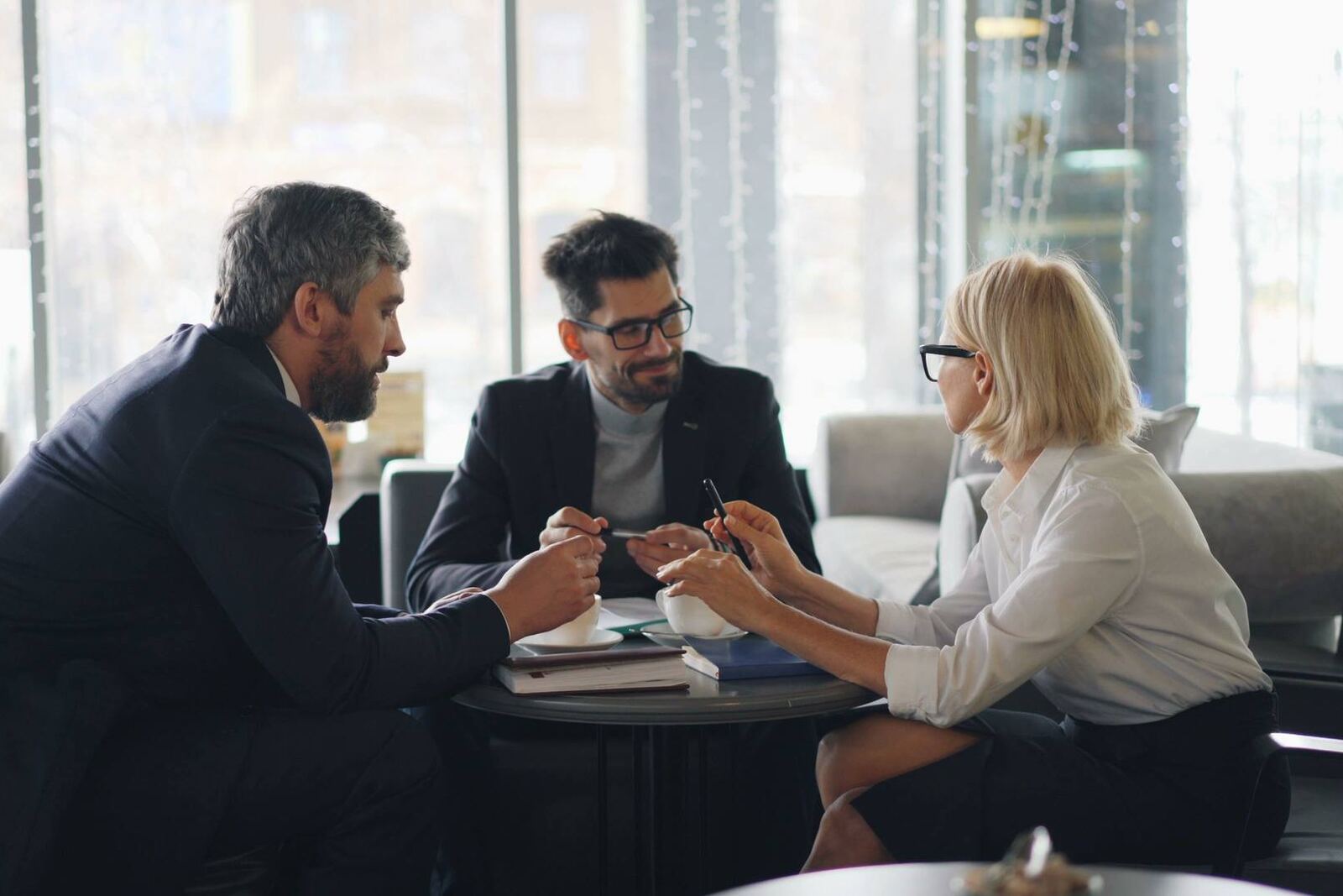 Three people in business attire lean in around a small table in a bright hotel lounge, mid-conversation with notebooks and coffee cups between them