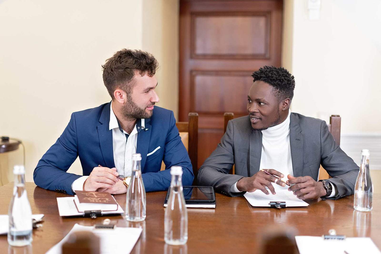 Two colleagues in suits sit at a wooden table lined with water bottles and papers, one gesturing mid-conversation beside a book titled The Law, a heavy wooden door behind them