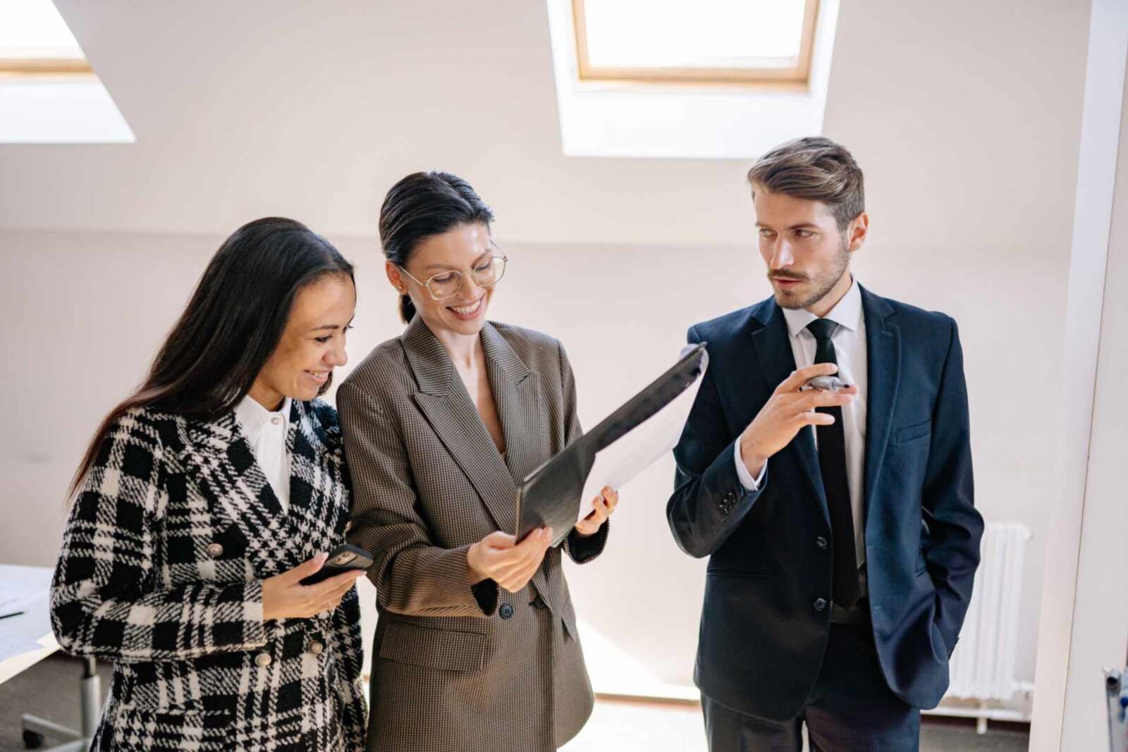 Two women in tailored jackets and a man in a suit and tie smile while reviewing a clipboard together in a bright room under skylights