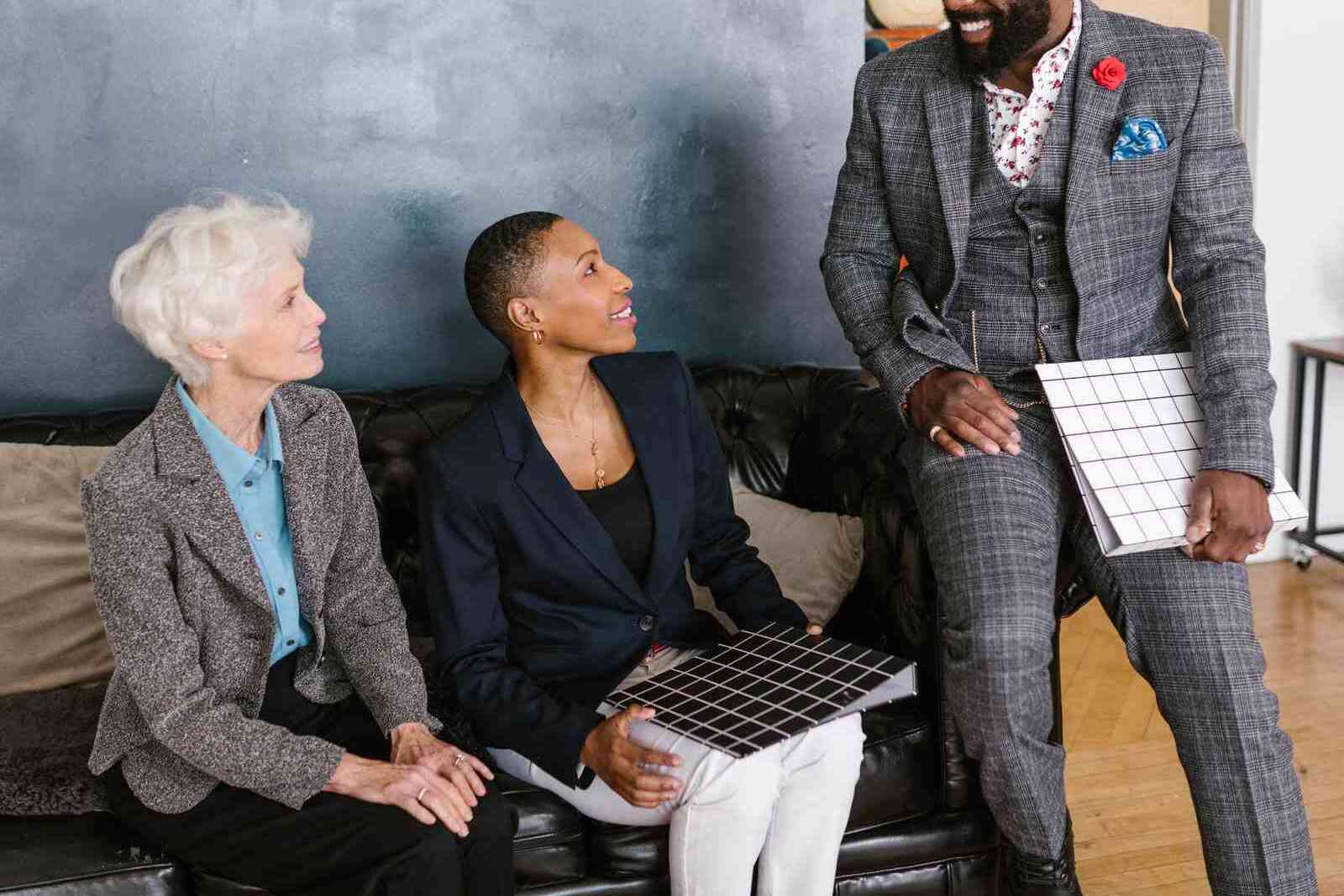 Three colleagues in business attire sit together on a leather sofa, laughing mid-conversation, each holding a gridded notebook